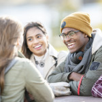 Five young adults are sitting at a picnic table outside on a fall day. They are smiling and chatting