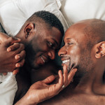 two black gay men holding hands and smiling in bed